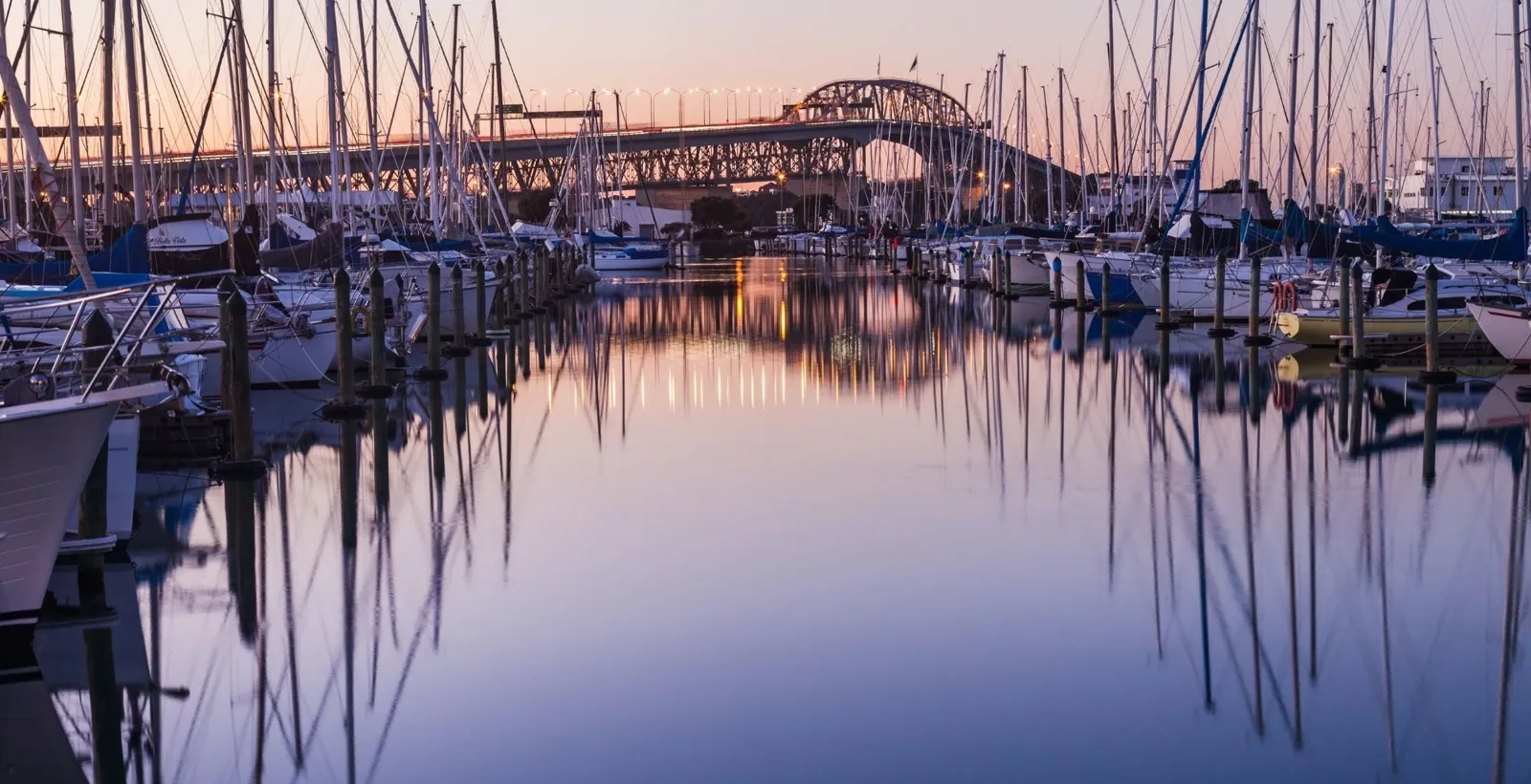 Yacht masts reflected in still marina water at dusk, with the Harbour Bridge behind