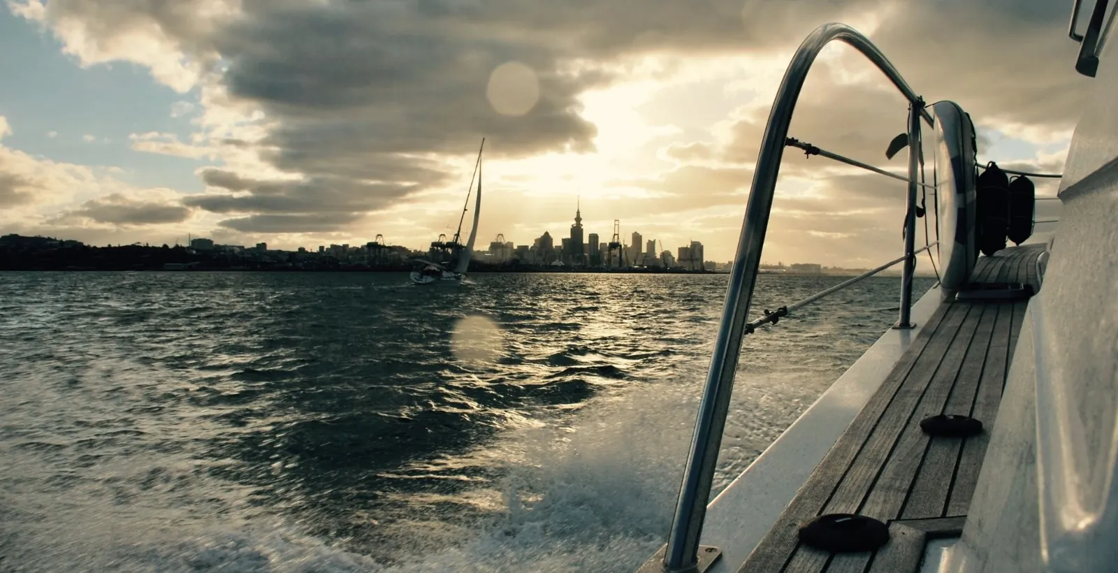 A yacht heeling in the wake beside a boat's rail, with the city skyline on the horizon