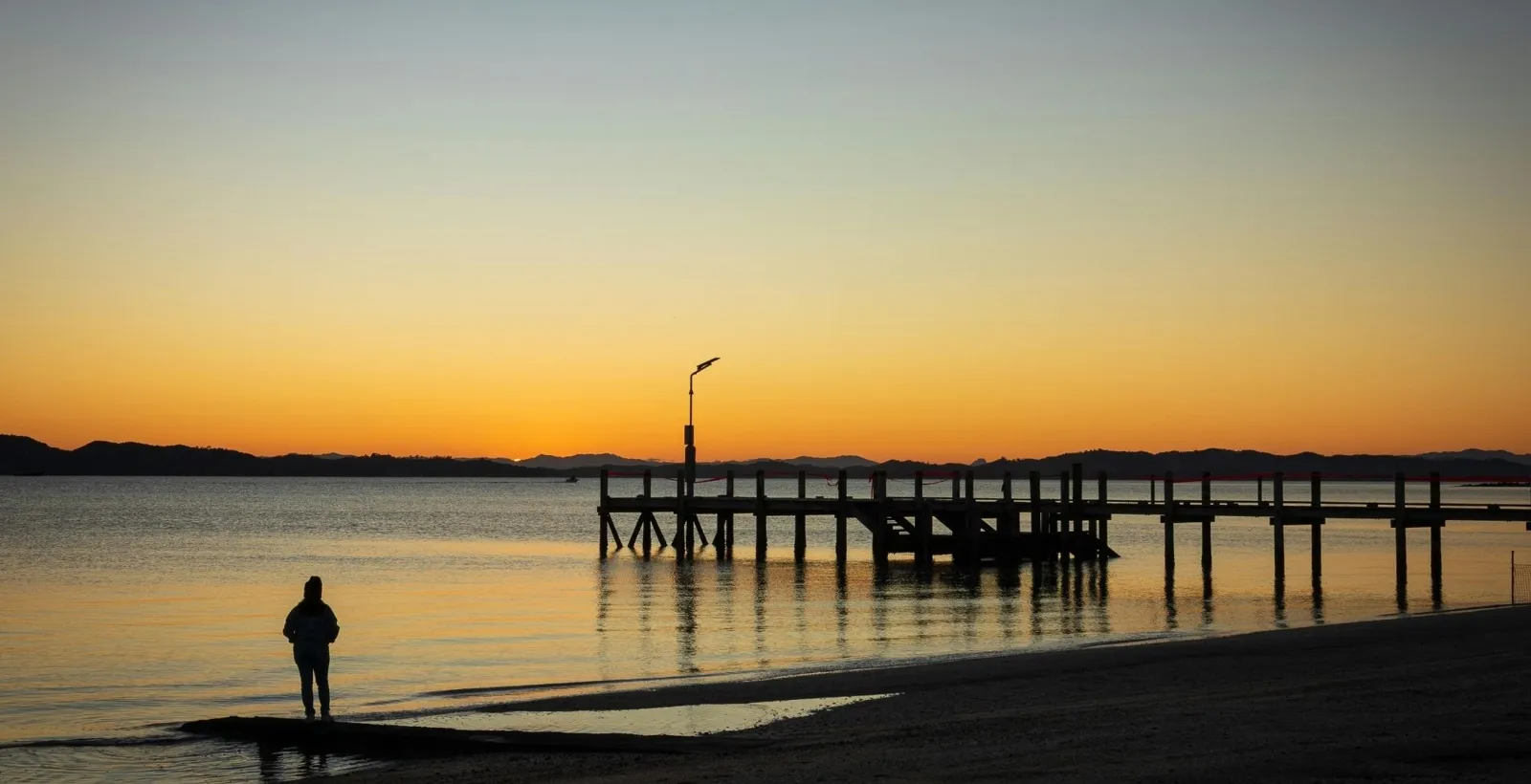 A person standing on the beach beside an old wooden jetty at sunset