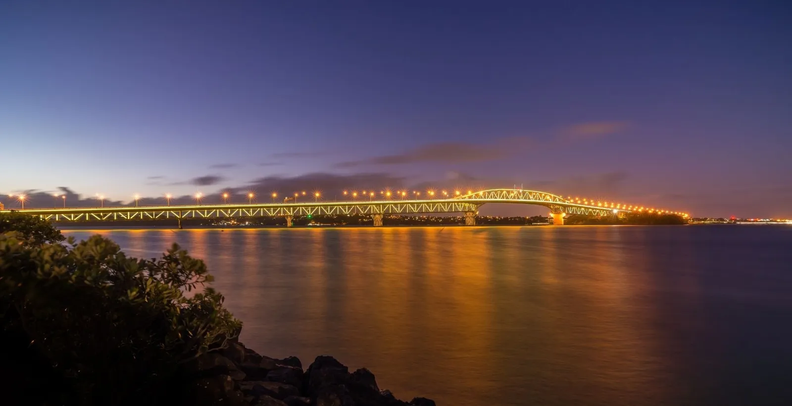 Auckland Harbour Bridge lit up at night, reflected in the harbour below