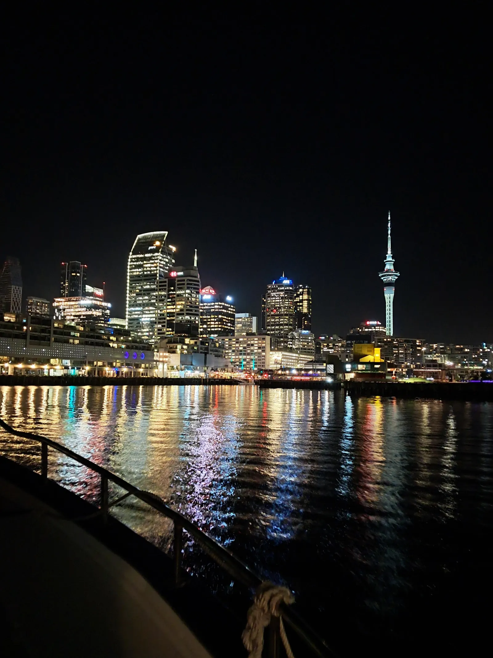 Night view of the lit waterfront and Sky Tower from beside a boat's deck rail