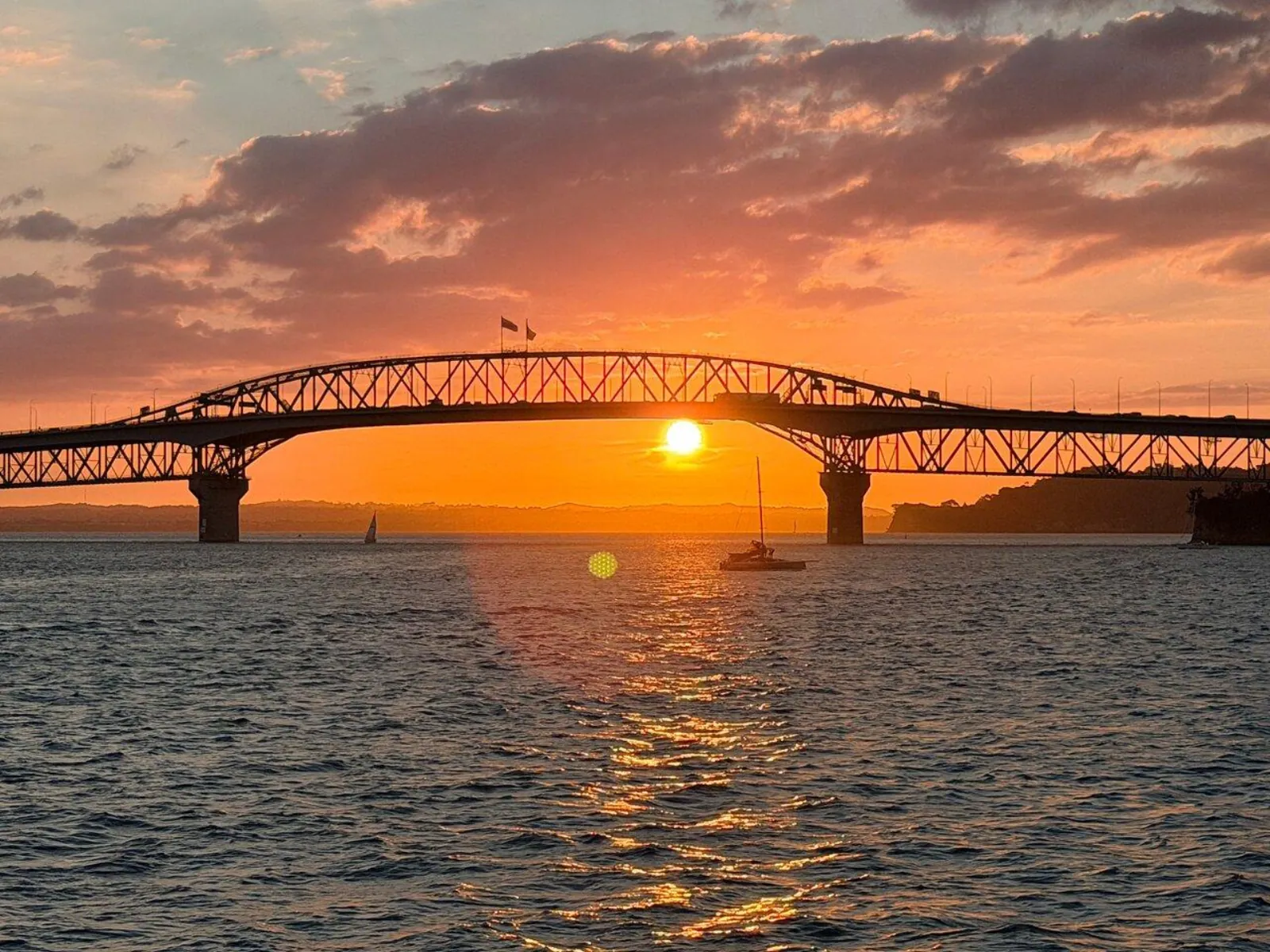 Sun sinking under the Auckland Harbour Bridge arch with a yacht in the golden glow