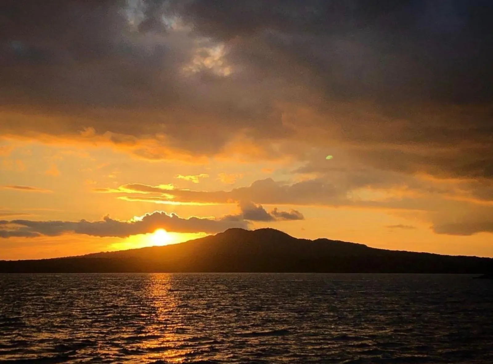 Low sun glowing beside the sloping silhouette of Rangitoto Island on the Waitematā