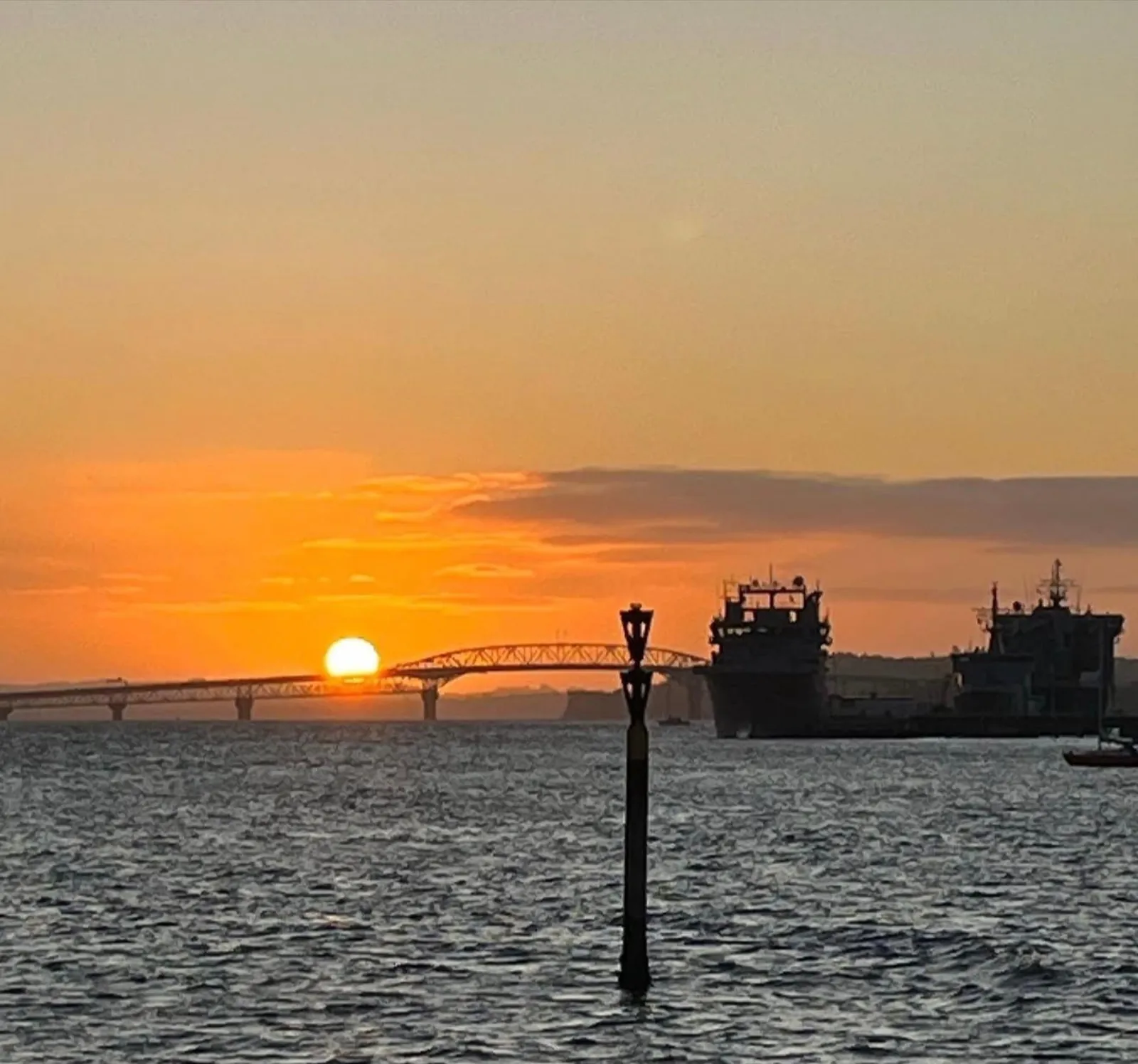 Sun dropping onto the Harbour Bridge beside moored ships and a channel marker post
