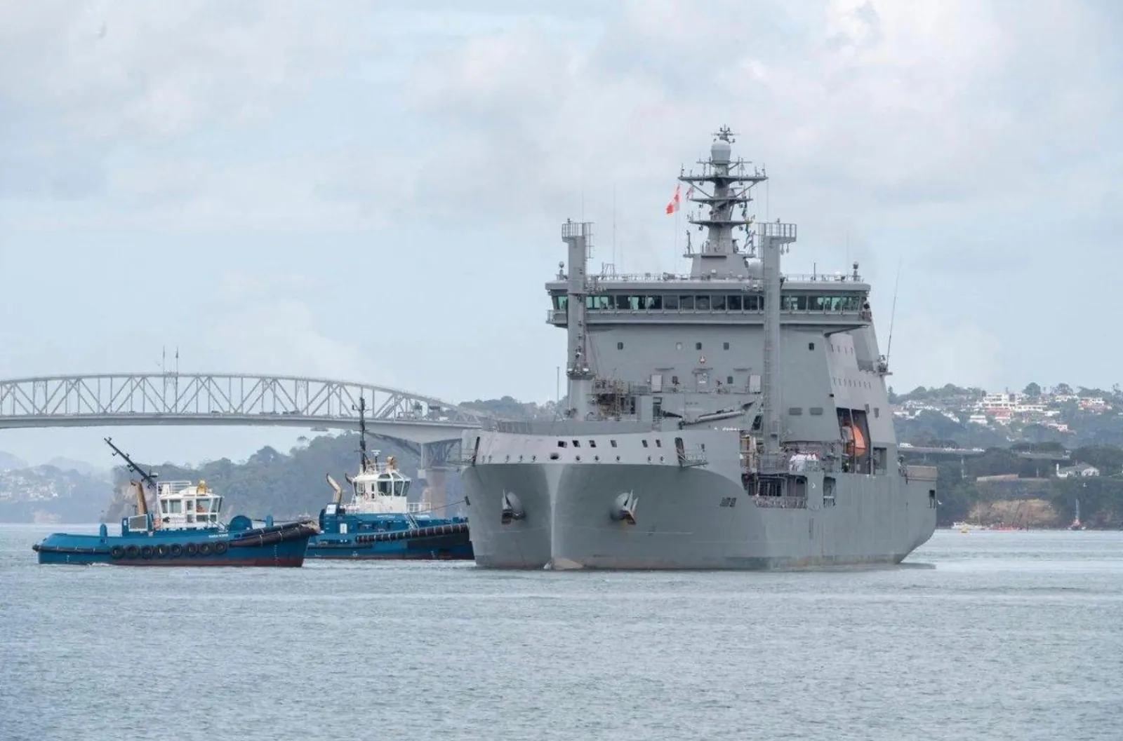 Grey naval ship escorted by tugboats in front of the Harbour Bridge