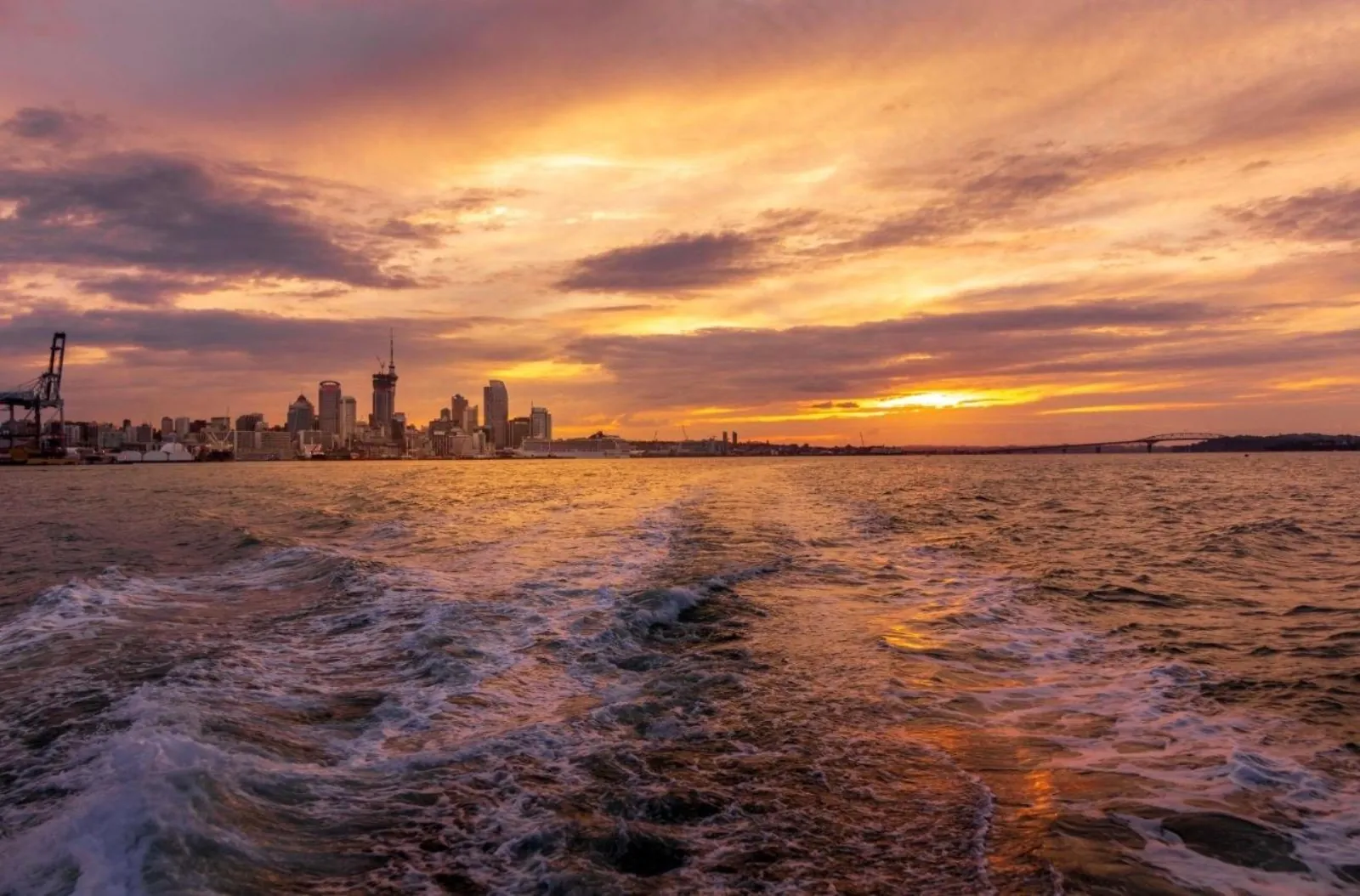 Boat wake trailing toward the Auckland skyline under an orange sunset sky