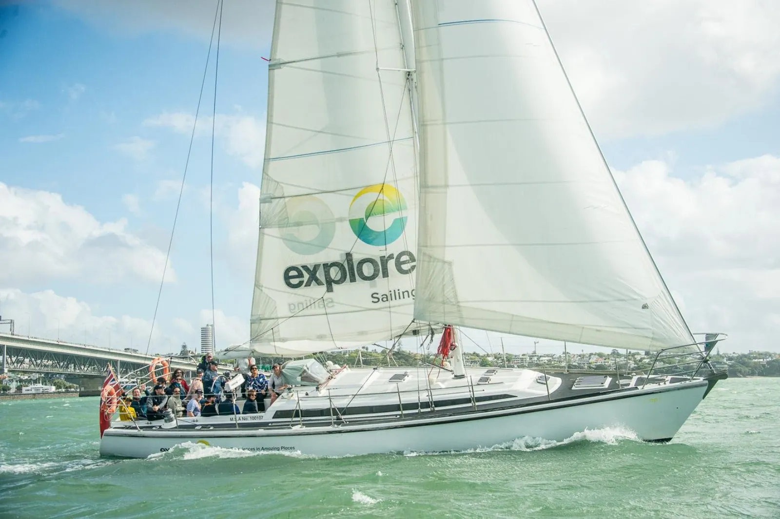 Cruising yacht with Explore sails passing the Harbour Bridge with guests on deck
