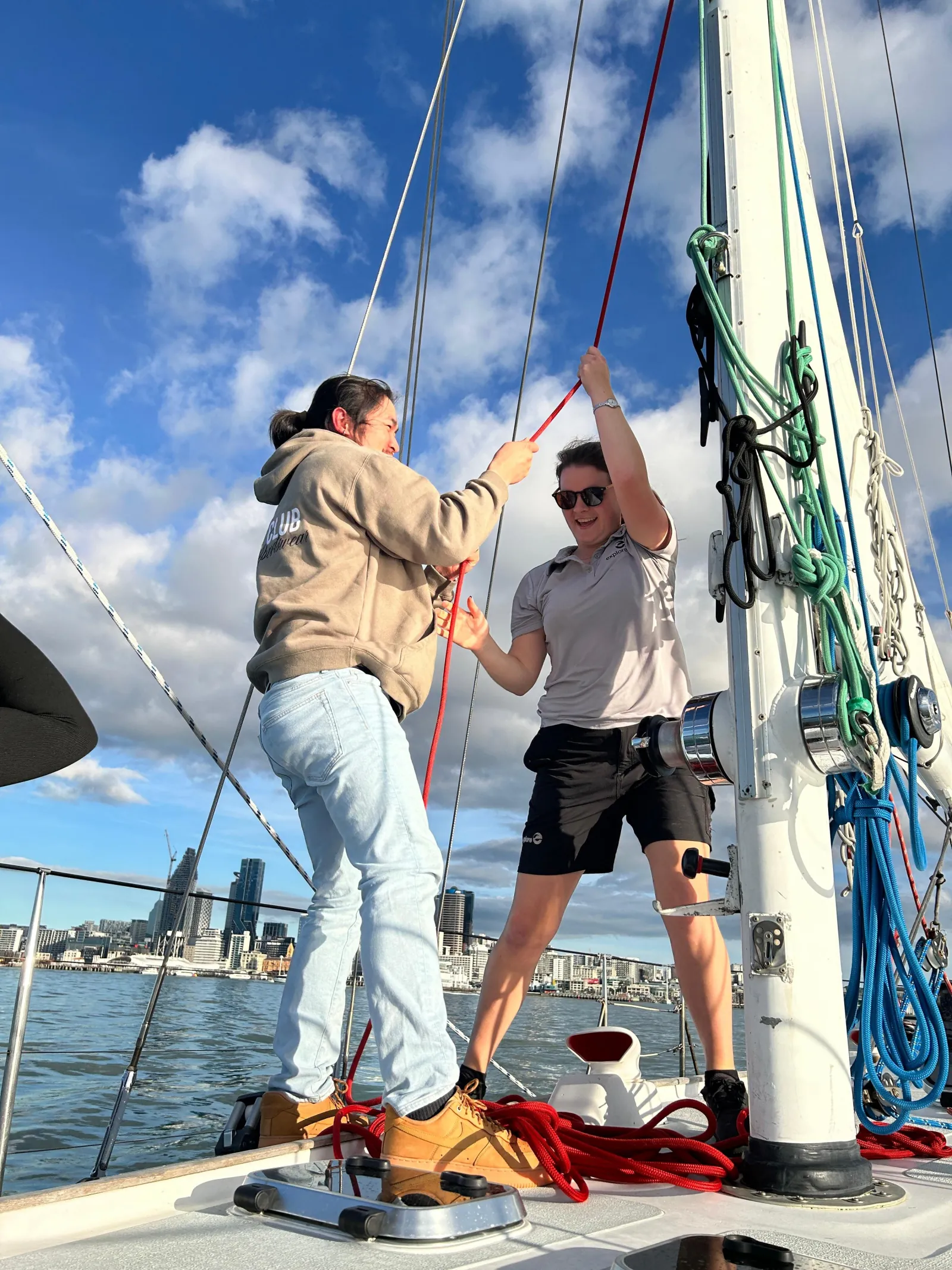 Two people hauling a halyard at the mast with the city behind